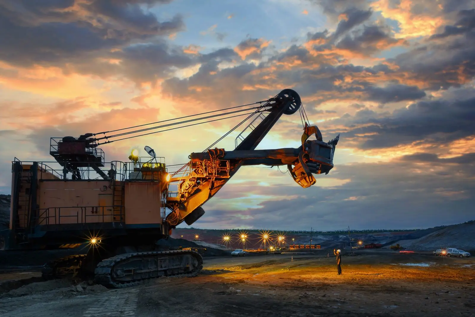 Giant mining shovel with its bucket raised against an orange dawn sky, site lights glowing and a worker standing nearby